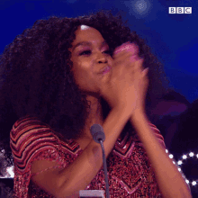 a woman with curly hair is clapping her hands in front of a microphone with the bbc logo in the corner