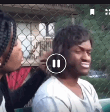 a woman is brushing a man 's hair in front of a chain link fence while playing a video .