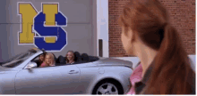 a group of girls are sitting in a silver convertible car in front of a building with the letter s on it .