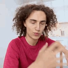 a young man with curly hair is sitting at a table with his eyes closed and a red shirt on .