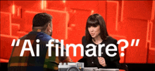 a man and a woman are sitting at a table and the woman is asking the man if he is filming