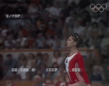 a female gymnast performs on a balance beam in front of a crowd with the olympics logo in the background