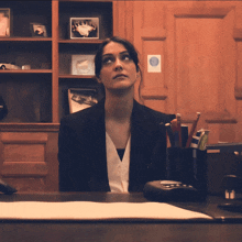 a woman in a suit sits at a desk in front of a wall with pictures on it