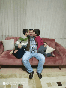 a man sits on a pink couch with two children kissing him on the cheeks