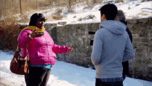 a woman in a pink jacket is talking to a man in a gray sweatshirt while standing in the snow .