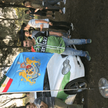 a man standing next to a car with a flag and a shirt that says lzz