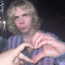 a person making a heart shape with their fingers in front of a man with blonde hair