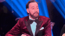 a man with a beard wearing a red tuxedo and bow tie is sitting in front of a blue curtain .