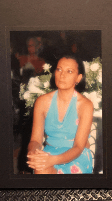 a woman in a blue dress sits with her hands folded in front of flowers