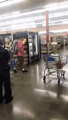 a man in a red jacket is pushing a shopping cart in a grocery store while a police officer watches