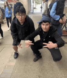 two young men are posing for a picture on a sidewalk . one is wearing glasses and the other is wearing a hat .