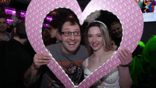 a man and a woman are posing for a picture behind a pink heart