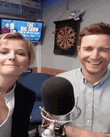 a man and a woman are smiling in front of a microphone with a dart board in the background that says the big show