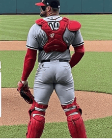 a baseball catcher is standing on a baseball field wearing a red vest and knee pads .