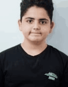 a young boy wearing a black t-shirt is standing in front of a white wall and looking at the camera .