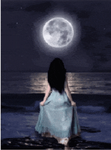 a woman in a blue dress is standing on the beach looking at the moon