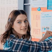 a woman in a plaid shirt is standing in front of a bulletin board that says " i am a poet "