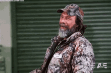 a man with a beard wearing a camouflage jacket and hat is standing in front of a green garage door .