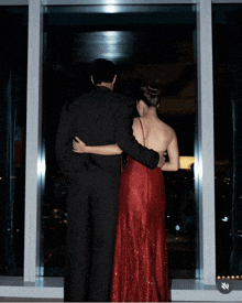 a man in a black suit and a woman in a red dress are looking out a window