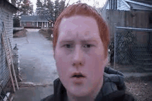 a young man with red hair is standing in front of a chain link fence and a house .