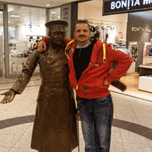 a man in a red jacket stands next to a statue in front of a store called bonita