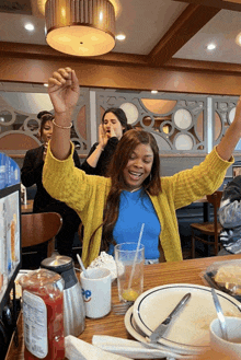 a woman in a yellow sweater is sitting at a diner table with her arms in the air