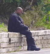 a bald man is sitting on a brick wall next to a road .