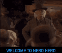 a man in a hat is standing in front of a crowd of people and says `` welcome to nerd herd '' .