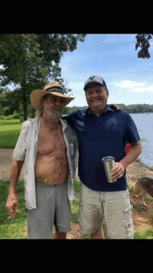 two men are posing for a picture in front of a body of water . one of the men is wearing a cowboy hat .