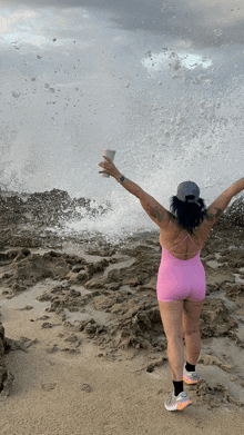 a woman in a pink bodysuit stands on a beach holding a cup