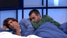 two men laying on a bed with one wearing a green sweater