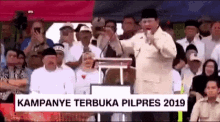 a man is giving a speech in front of a crowd while a sign says kampanye terbuka pilpres 2019 .