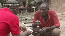 two men are sitting on the ground playing cards and one of them is holding a piece of paper in his hand .