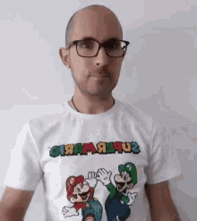 a bald man wearing glasses and a mario and luigi t-shirt is standing in front of a white wall .