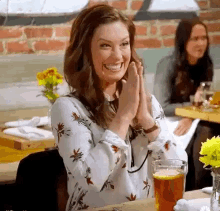 a woman is clapping her hands while sitting at a table with a glass of beer in front of her .