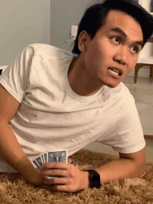 a man in a white t-shirt is laying on the floor holding playing cards