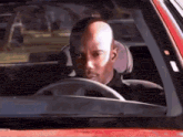 a bald man is driving a red car with his head out the window