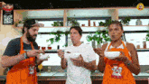 two men and a woman are eating from bowls in a kitchen with an apron that says ' ead ' on it