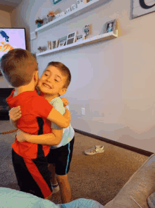 a boy in a red shirt is hugging another boy in a blue shirt in a living room