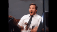 a man in a white shirt and tie is sitting at a table with his mouth open and a surprised look on his face .