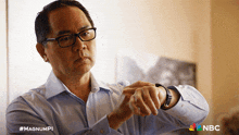 a man wearing glasses is looking at his watch with a nbc logo in the corner