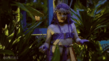 a woman with purple hair and a purple top is standing in a jungle .