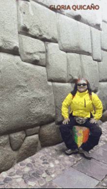 a woman in a yellow jacket is squatting down in front of a stone wall with the name gloria quicaño written on the bottom