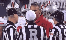 a group of football players are standing around a referee wearing a shirt that says ' ohio state ' on it .