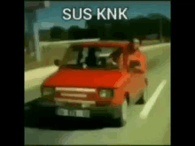 a man is driving a red car down a highway with a caption that says sus knk .