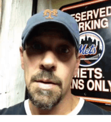 a man wearing a mets hat stands in front of a served parking sign