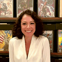 a woman in a white shirt is smiling in front of a shelf of comic books including one called iron man