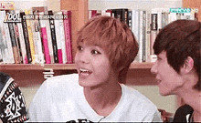 two young men are sitting next to each other in front of a bookshelf and talking to each other .