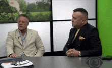 two men are sitting at a table with their hands folded in front of a green wall