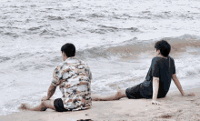 two men are sitting on the beach looking at the water